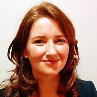 Woman with light skin and auburn hair smiles at the camera, wearing a dark jacket.