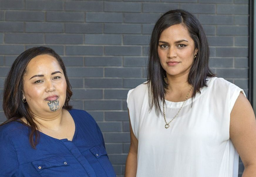 Two women stand in front of a gray brick wall. One wears a blue shirt and has a facial tattoo, the other a white top.