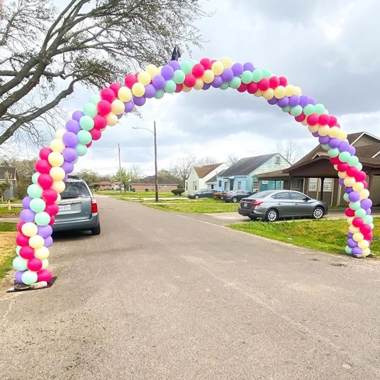 Balloon arch of pastel colors in a residential street. A car is parked on the road under the arch.