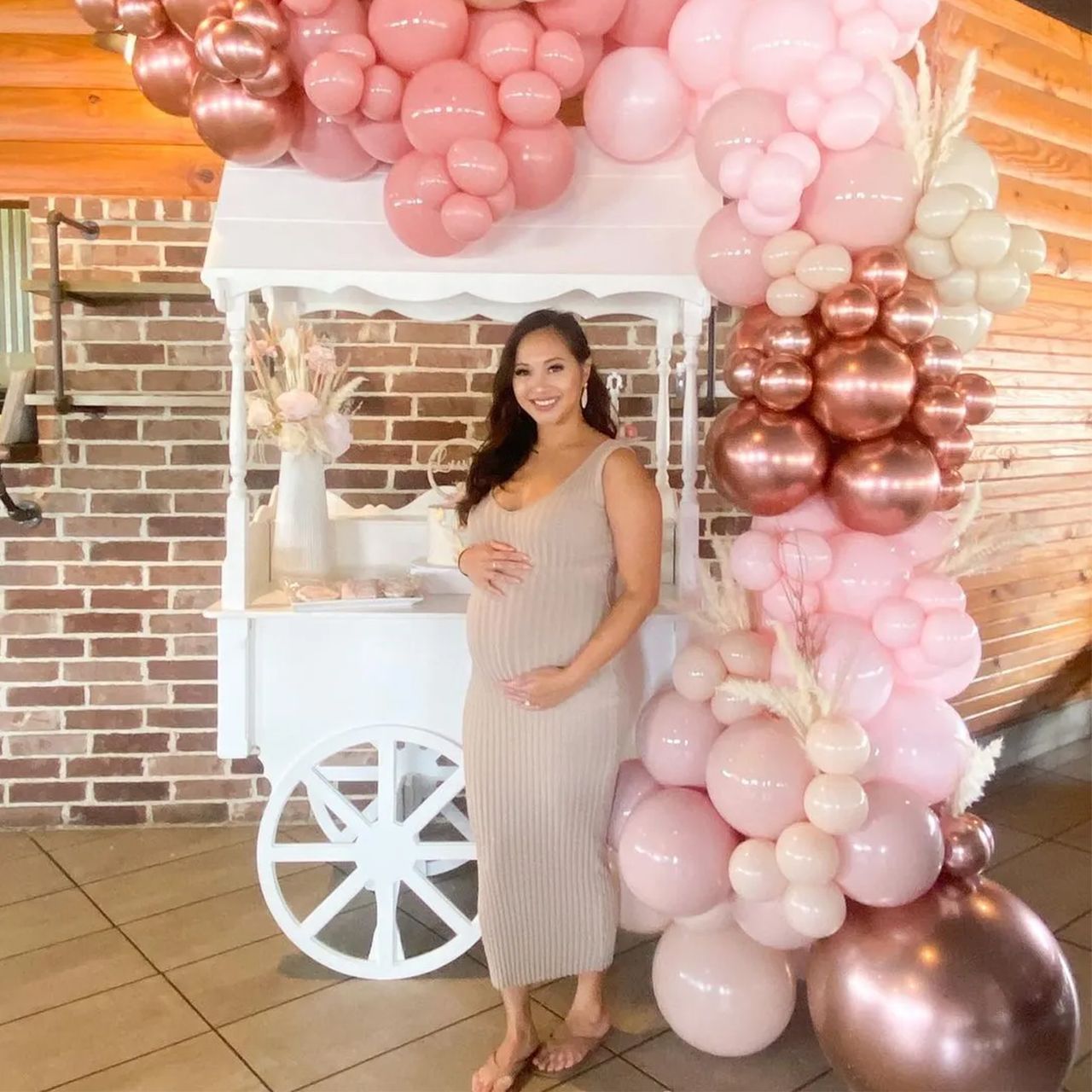 Pregnant person in beige dress at a party, holding belly. Decorated with pink and copper balloons and a white cart.