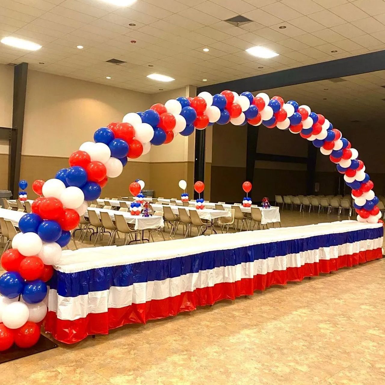 Arch of red, white, and blue balloons over a table decorated similarly. Indoor event setting with tables and chairs.