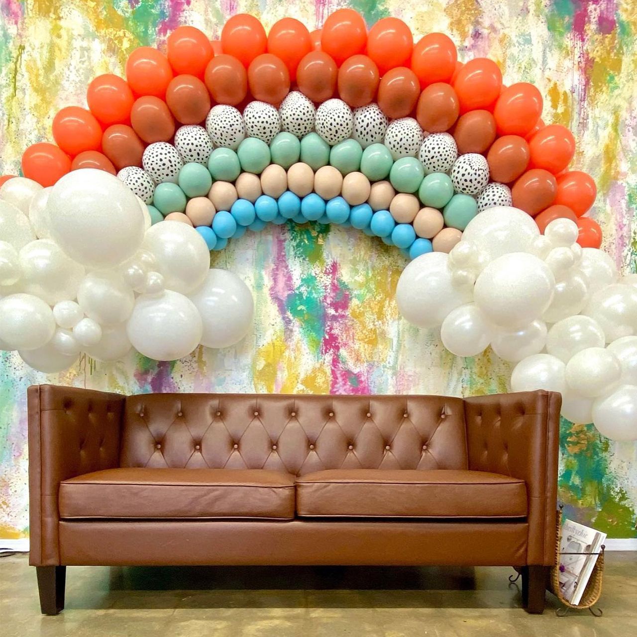 Brown sofa below a rainbow balloon arch against a colorful wall; white balloon clouds on the sides. Brown sofa below a rainbow balloon arch against a colorful wall; white balloon clouds on the sides.