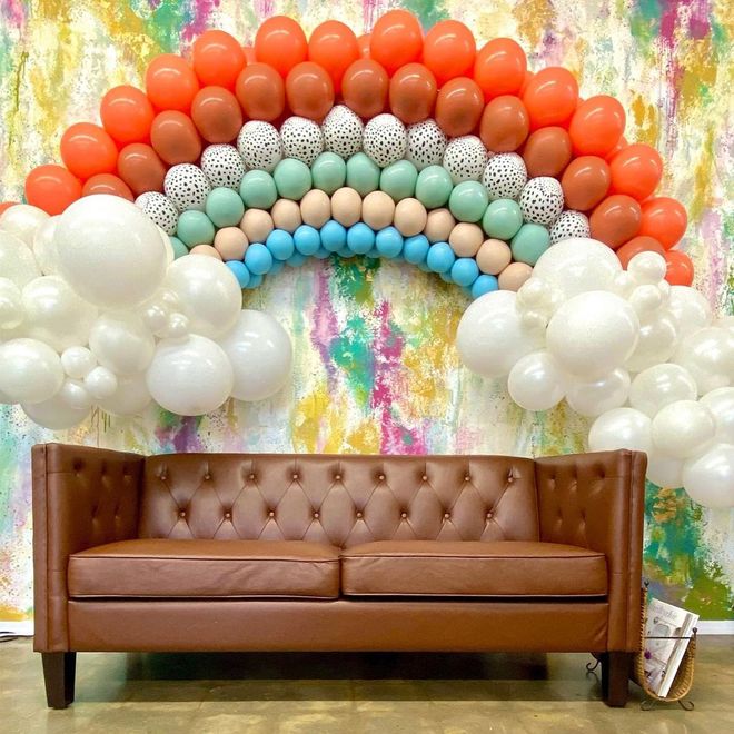Brown sofa below a rainbow balloon arch against a colorful wall; white balloon clouds on the sides. Brown sofa below a rainbow balloon arch against a colorful wall; white balloon clouds on the sides.