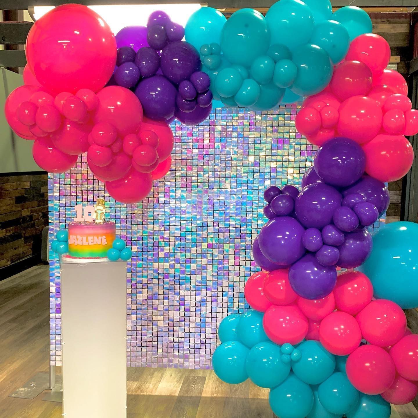 Party scene: balloons (pink, purple, teal) surround a shiny square backdrop and cake on a pedestal.