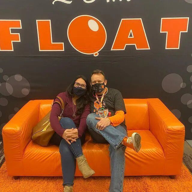 Two people wearing masks sit on an orange couch in front of a