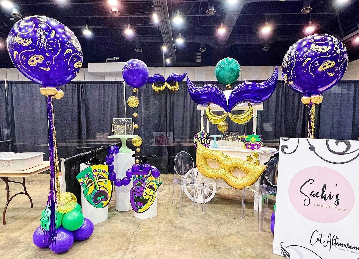 Mardi Gras-themed display: purple and gold balloons, mask decorations, a cart, and sign.