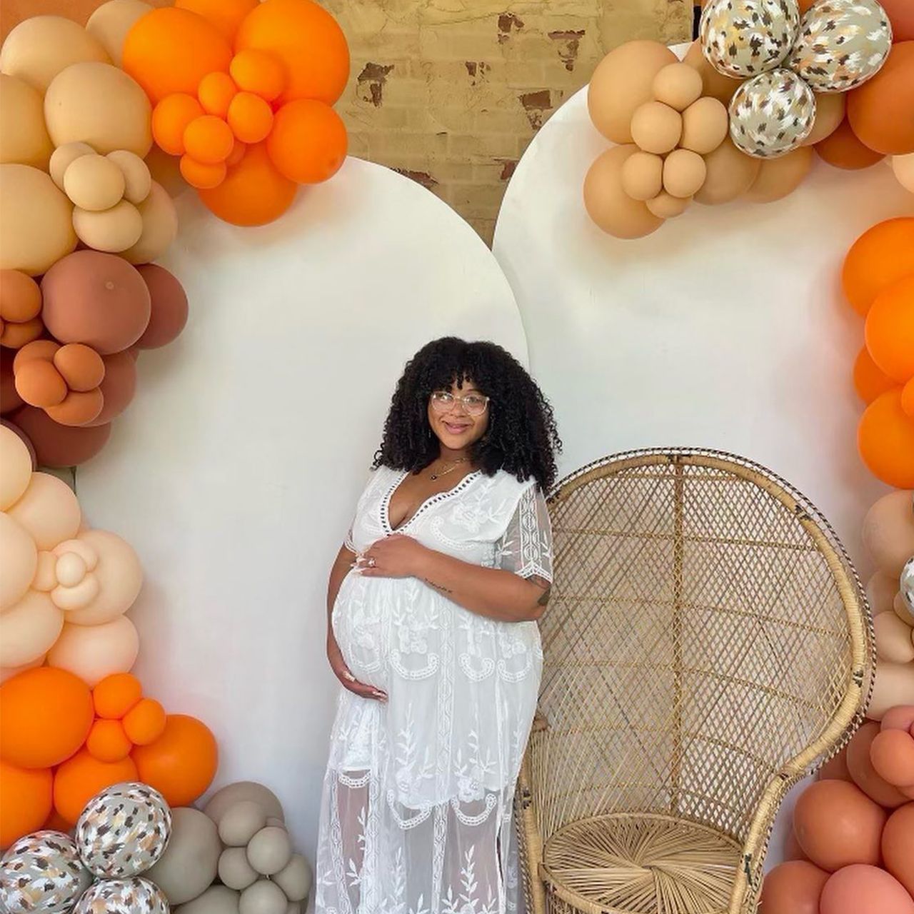 Pregnant person in white dress with hands on belly, standing near a wicker chair and balloon arch in shades of orange and tan.