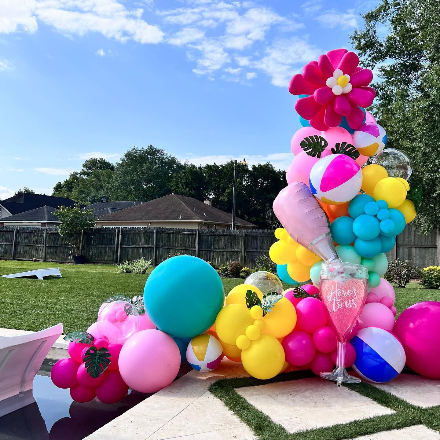Colorful balloon arch by a pool, featuring beach balls, flowers, and a clear cup. Sunny outdoors.