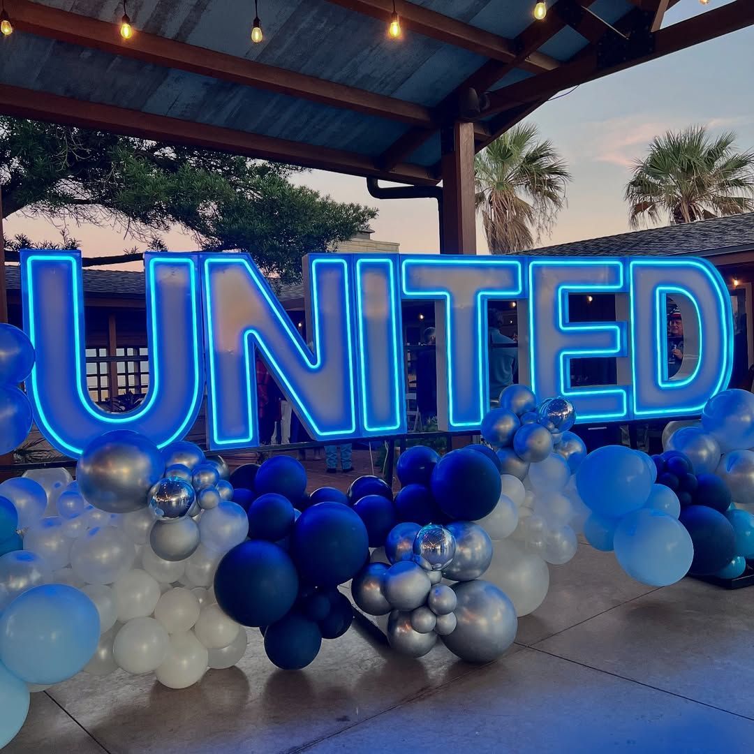 Woman in white dress stands near balloon arch and welcome sign, overlooking water.