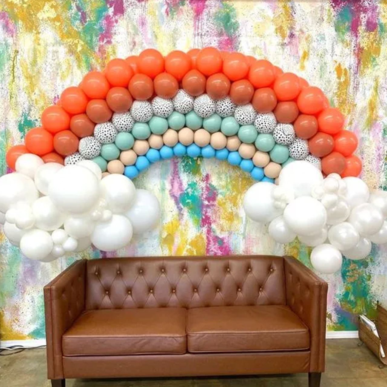 Rainbow balloon arch with white clouds above a brown leather couch against a colorful wall.