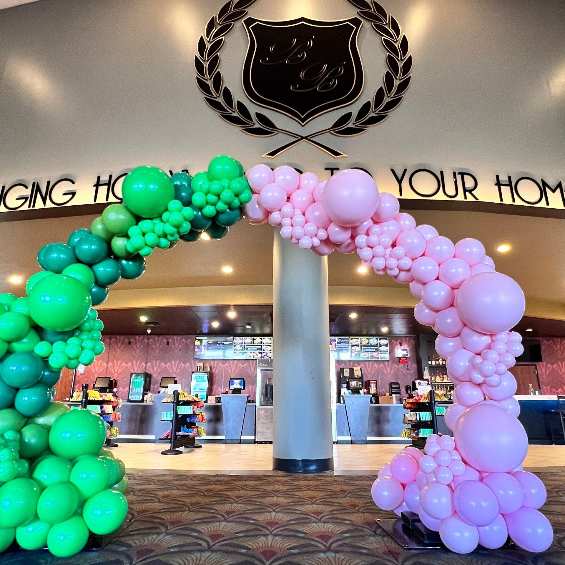 An archway of green and pink balloons at a venue entrance, with a crest above.