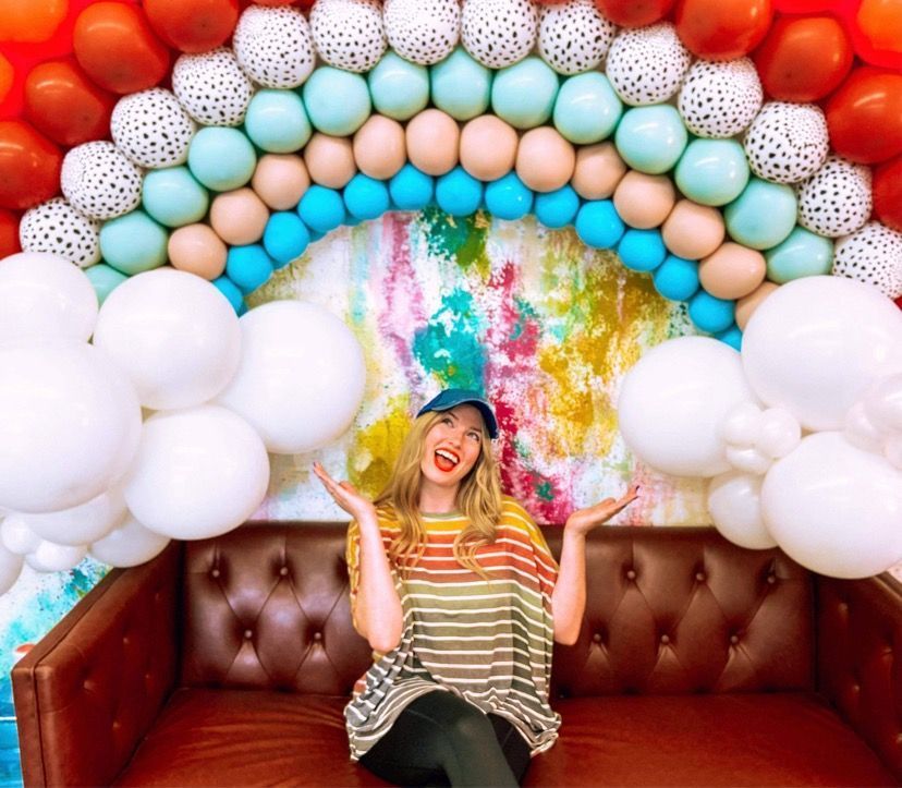 Woman sits on red couch, arms raised, smiling, in front of a colorful balloon archway.