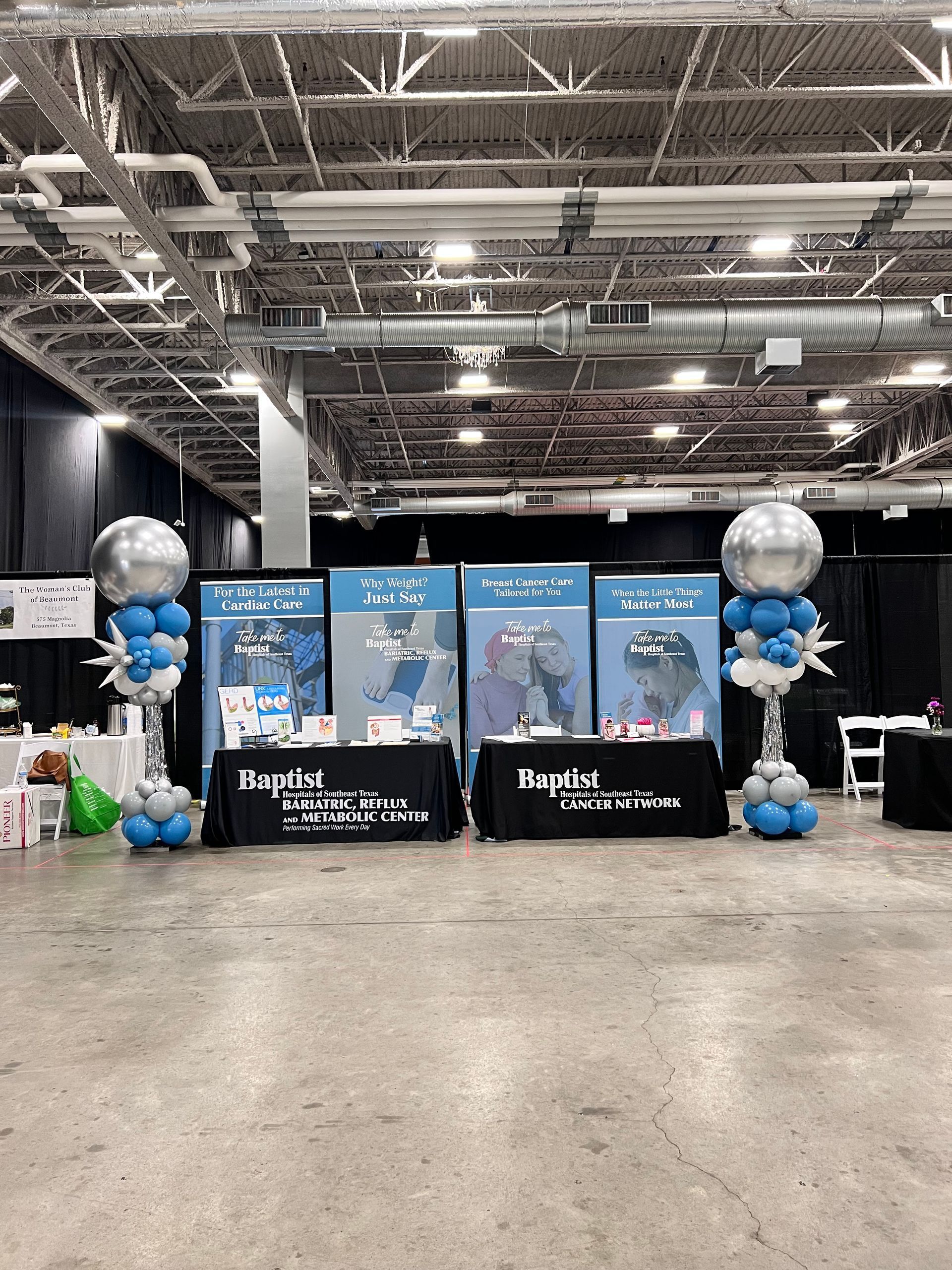 Booth with blue and white balloons, displays, and tables. Event setting with overhead lighting.