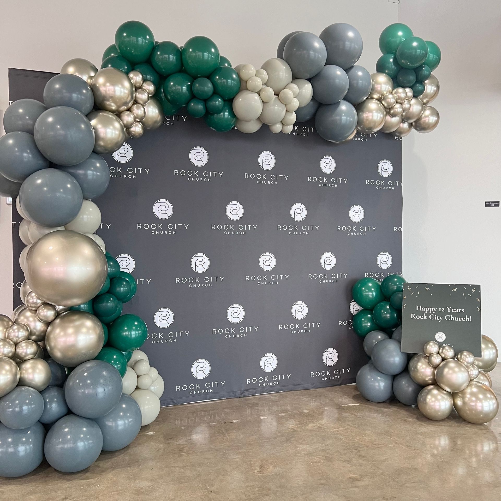 Dark backdrop with a balloon arch in shades of green, gray, and gold. Sign on the right.