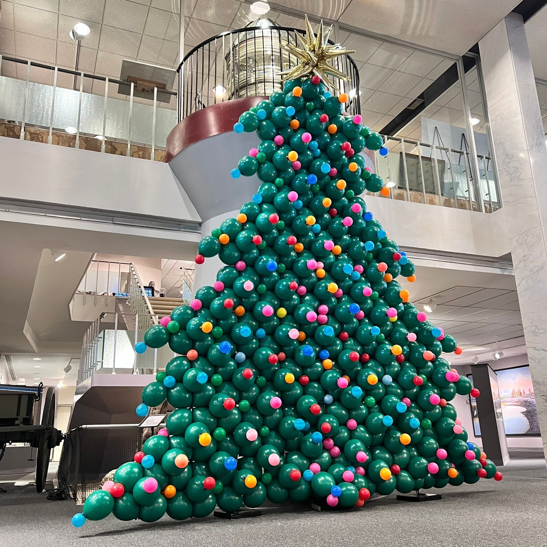 Christmas tree sculpture made of green balloons, with colorful ornaments, in a building with a spiral staircase.