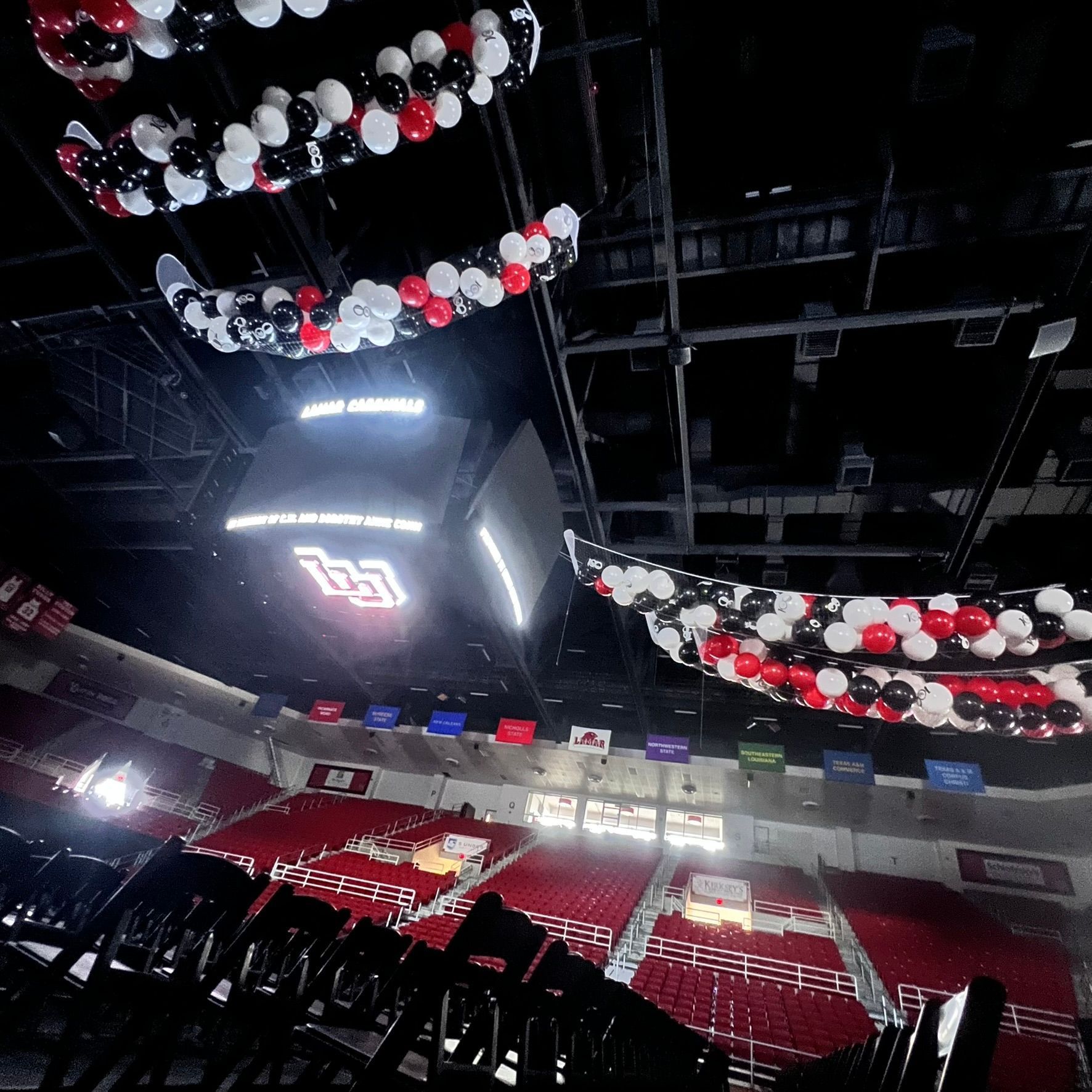 Arena interior with red seating, black chairs, and hanging balloons in red, white, and black. A scoreboard is visible.