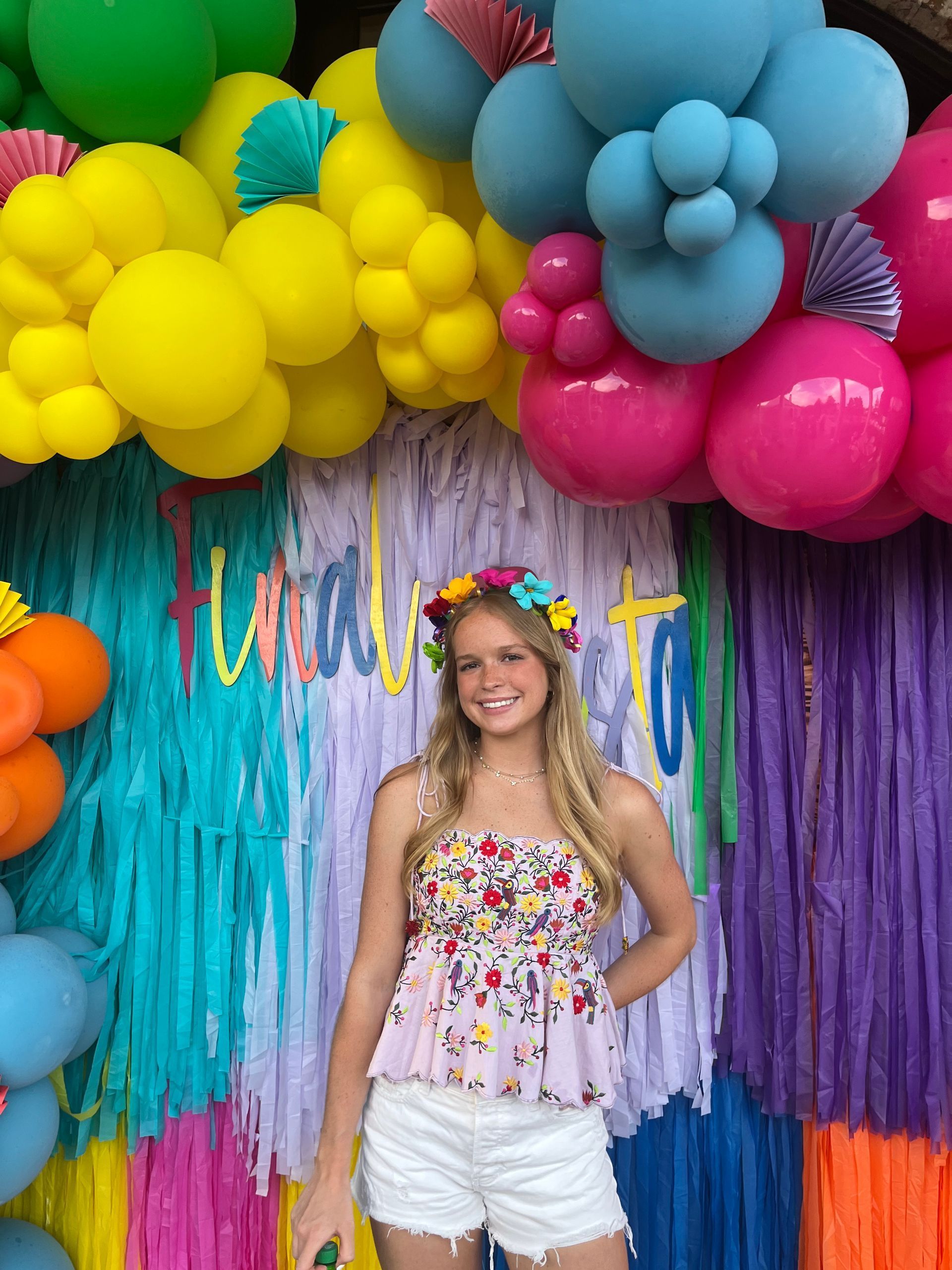 Young person smiles, wearing flower crown and floral top, in front of a colorful backdrop with balloons. Young person smiles, wearing flower crown and floral top, in front of a colorful backdrop with balloons.