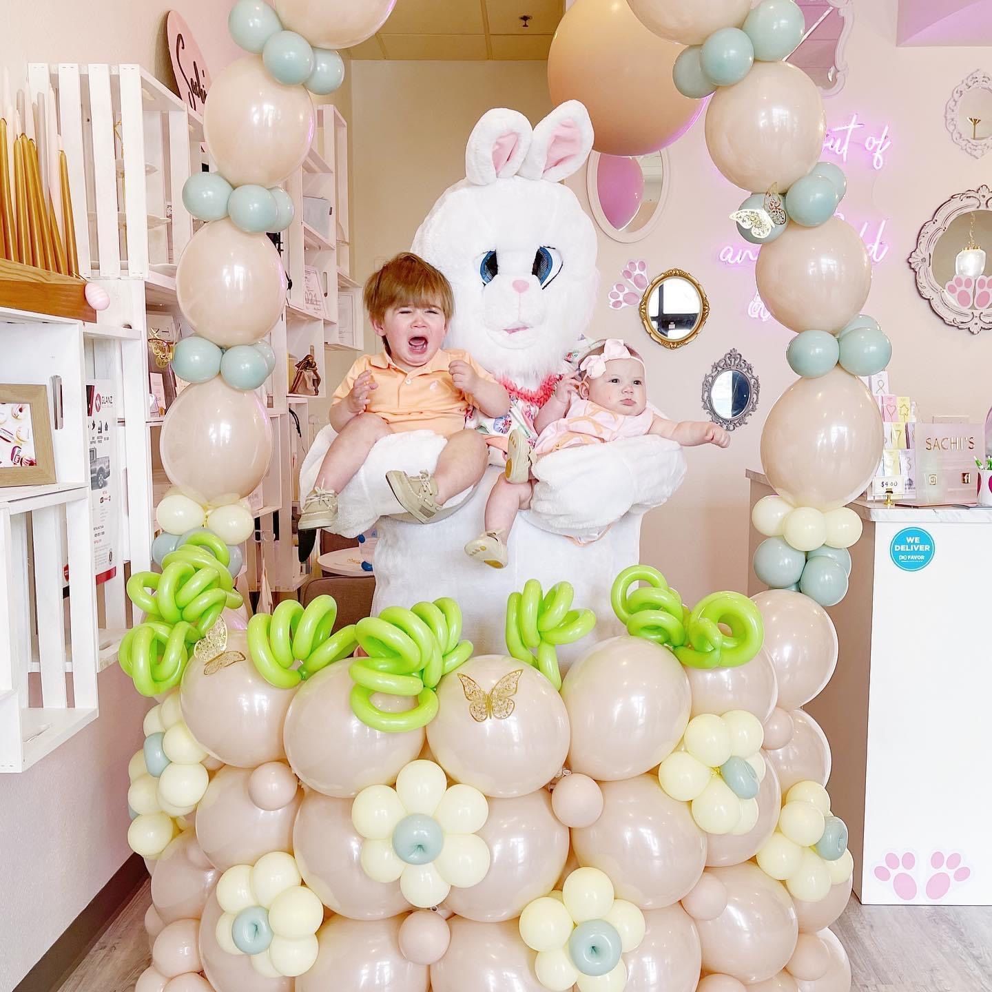 Two children held by a large bunny statue at a pastel-colored Easter balloon display.