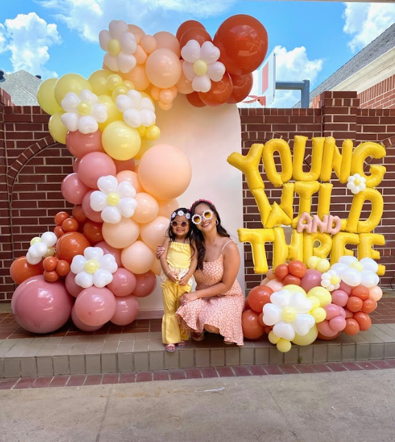 Mother and child pose in front of a “Young Wild and Three” balloon arch. Yellow, pink, orange balloons, brick background.