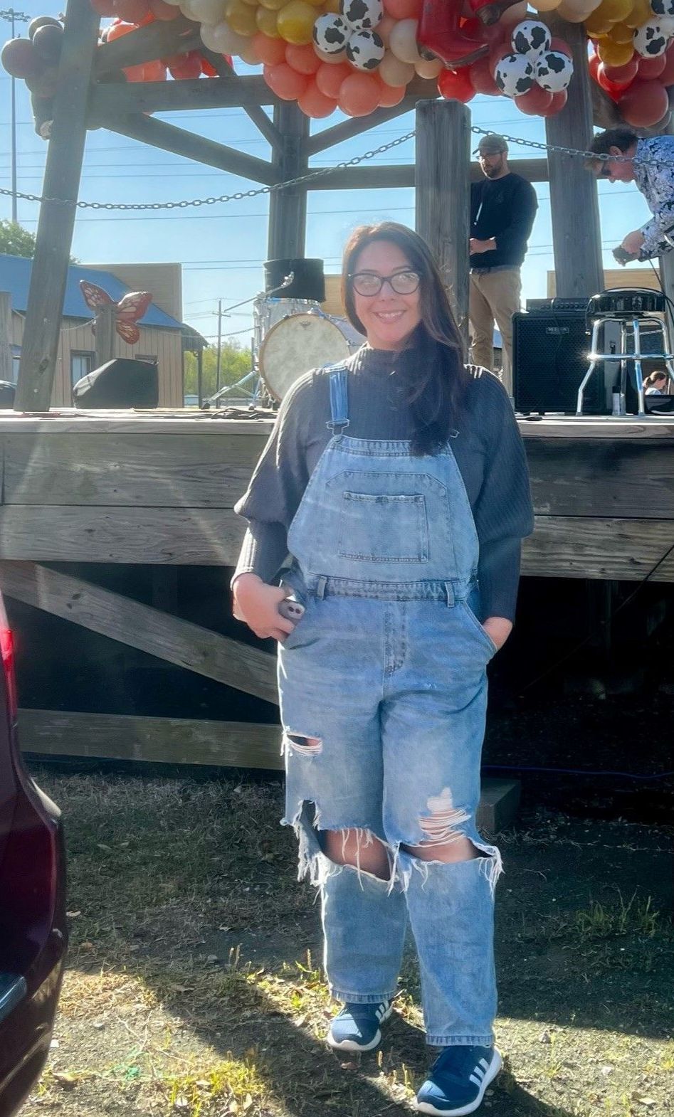 Woman in ripped overalls stands outside near a stage with balloons, wearing a gray sweater.