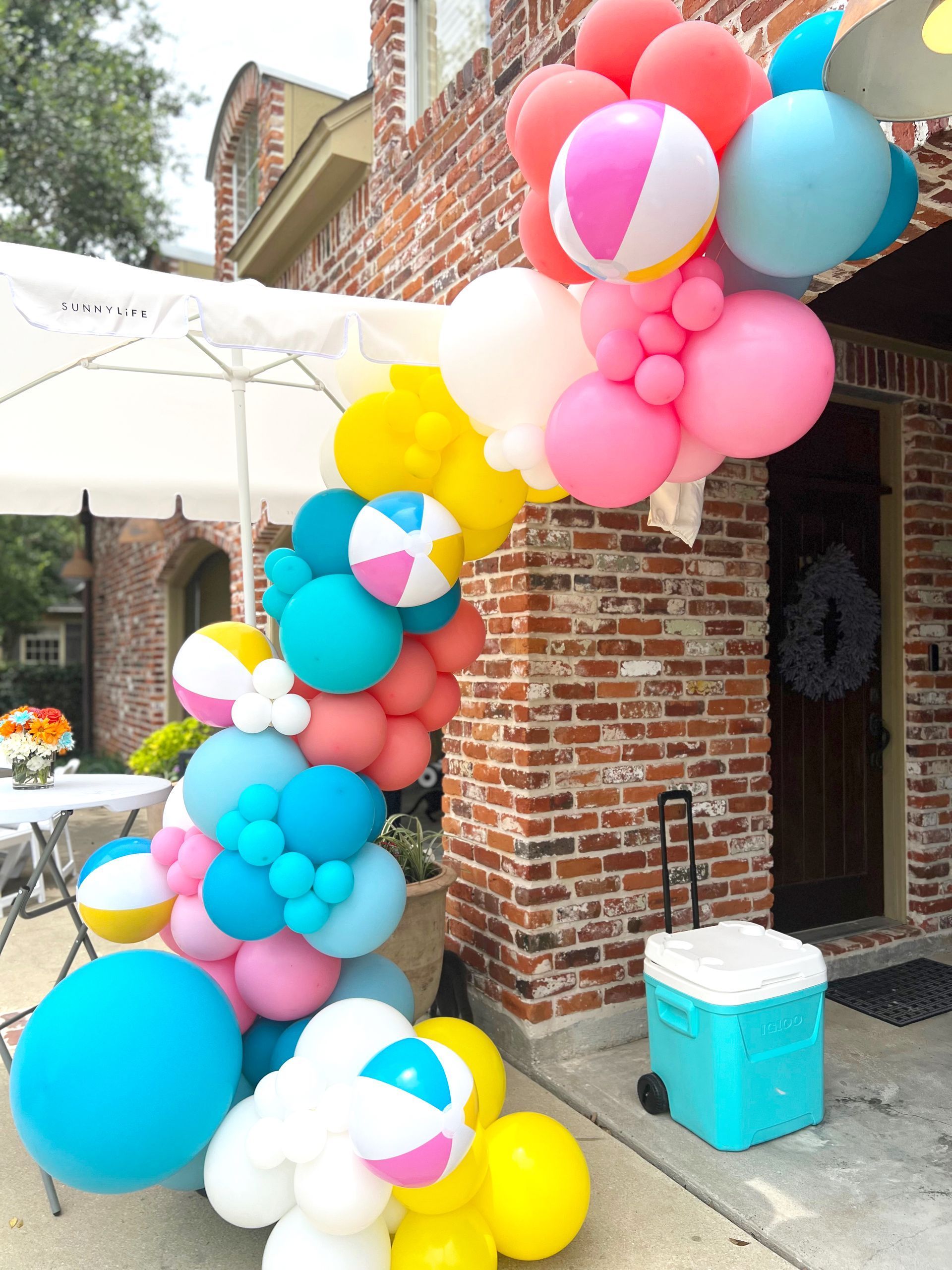 Colorful balloon arch with beach balls, against a brick building with a teal cooler beside it.