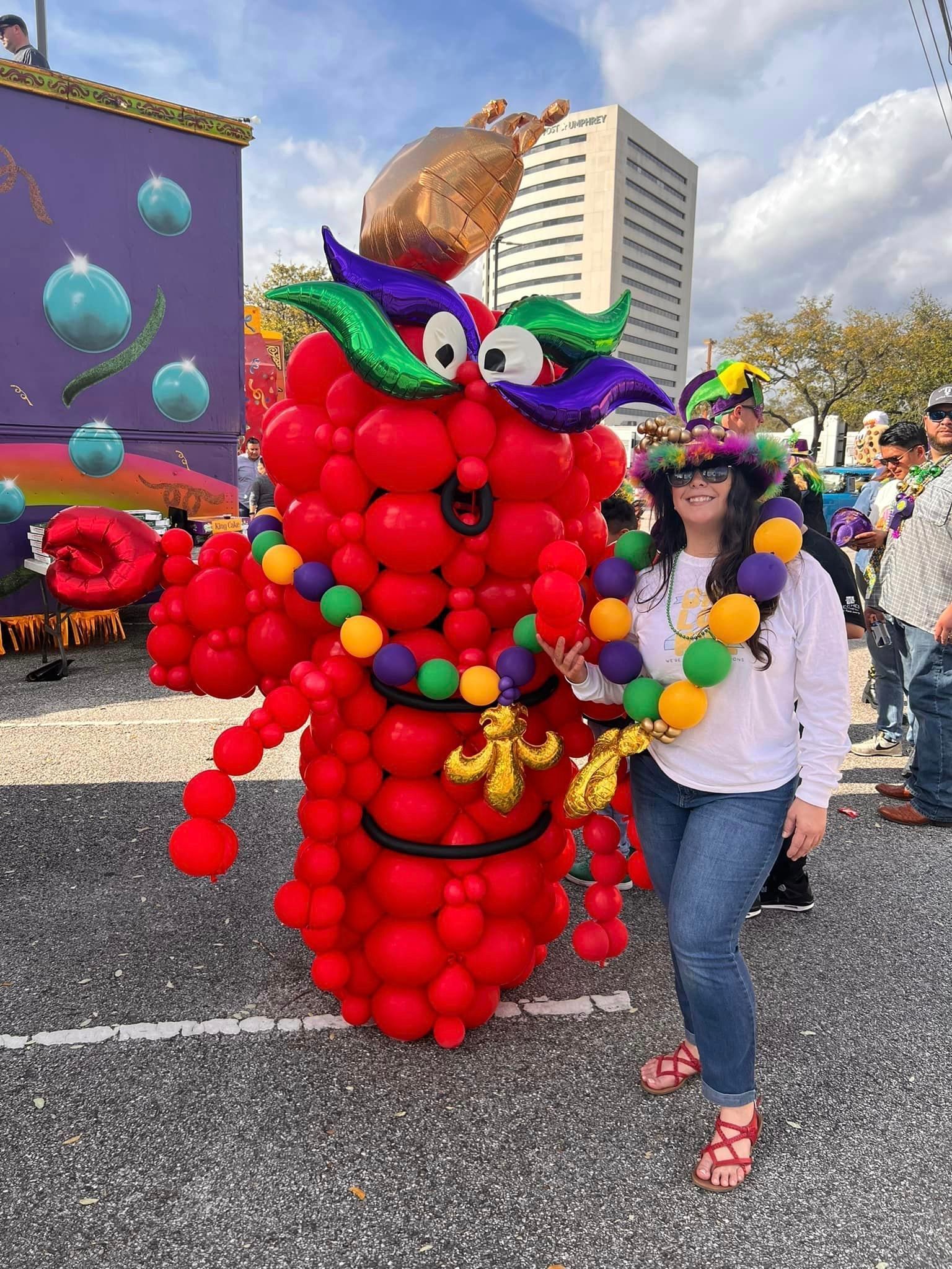 Woman in jeans and beads poses with a red balloon creature at a parade; a float is behind them.