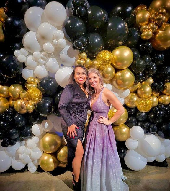 Two women pose in front of a balloon wall with black, white, and gold balloons.