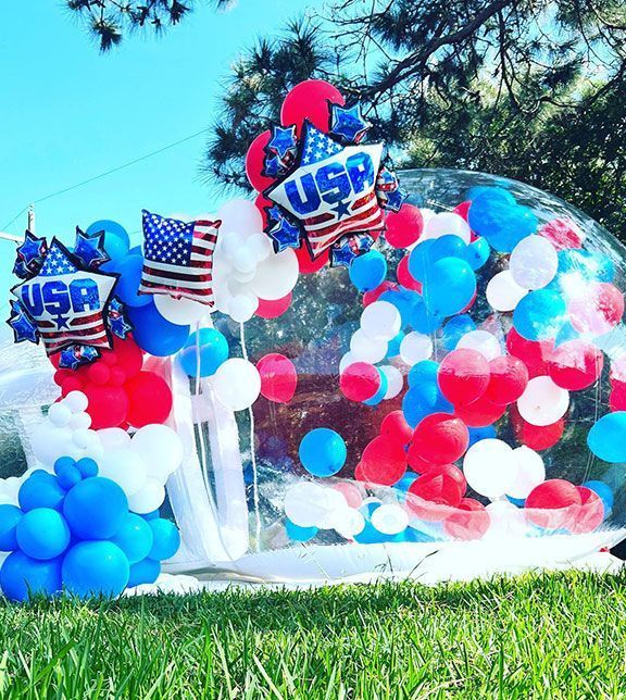 A clear bubble structure filled with red, white, and blue balloons, and American flag decorations on grass.