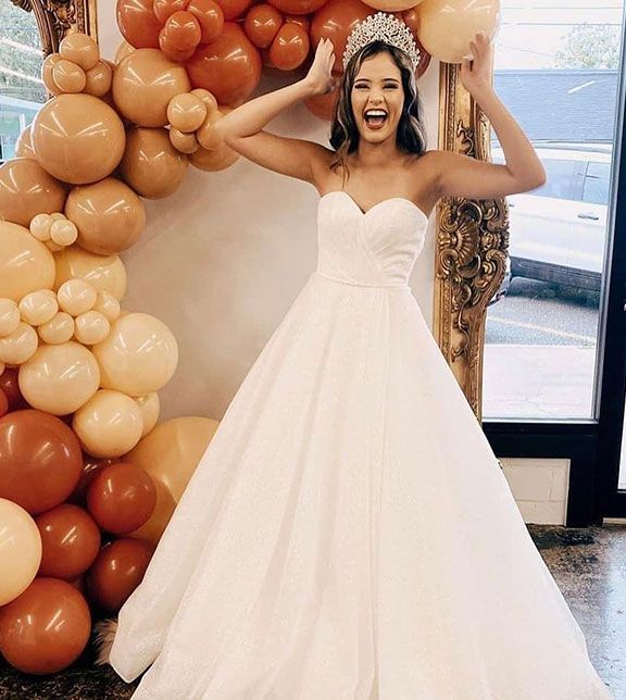 Woman in white gown and tiara, arms raised, smiling widely in front of a balloon arch.