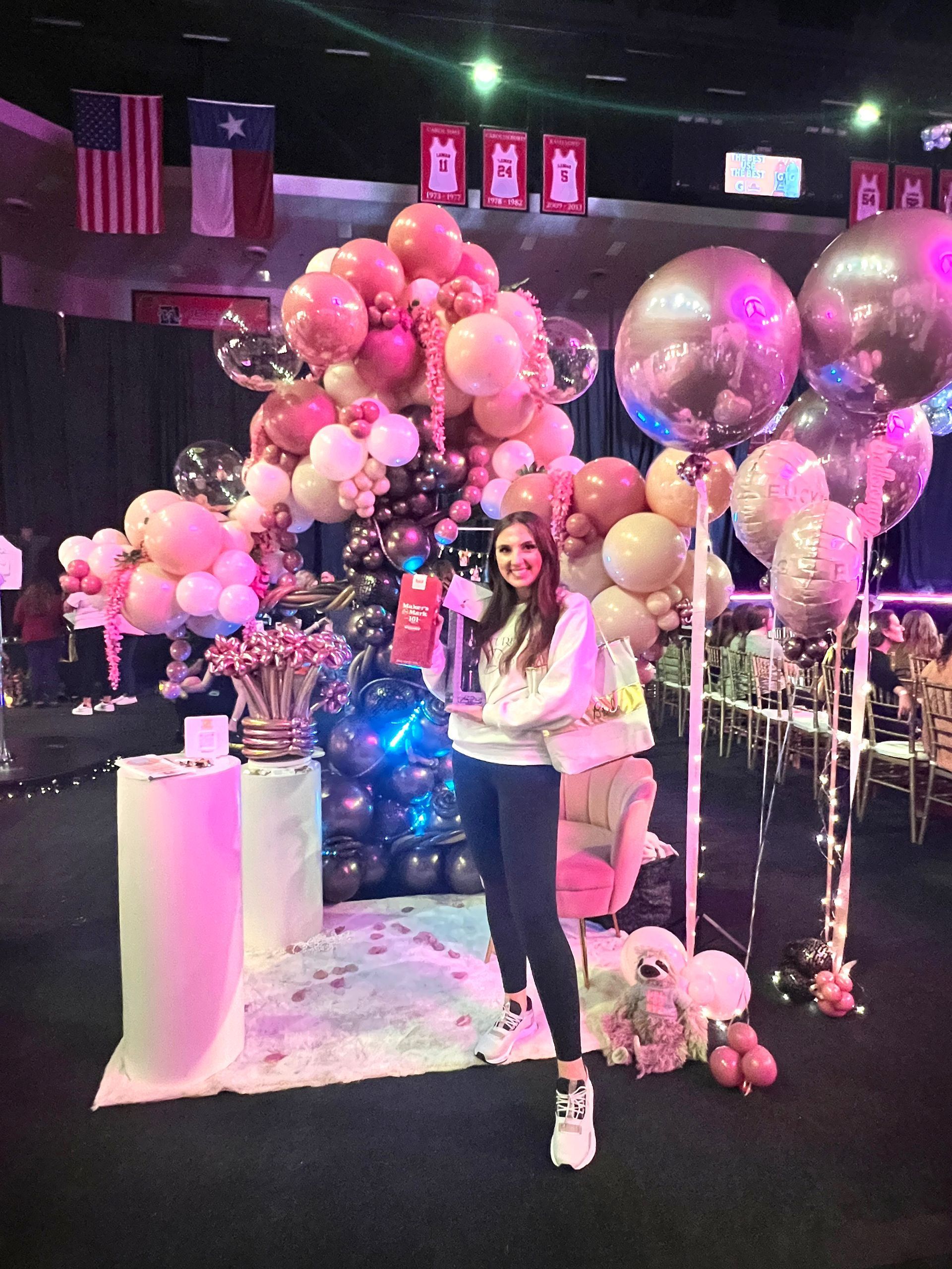 Woman posing with balloons at an event. Pink, gold, and black balloons, party setting.