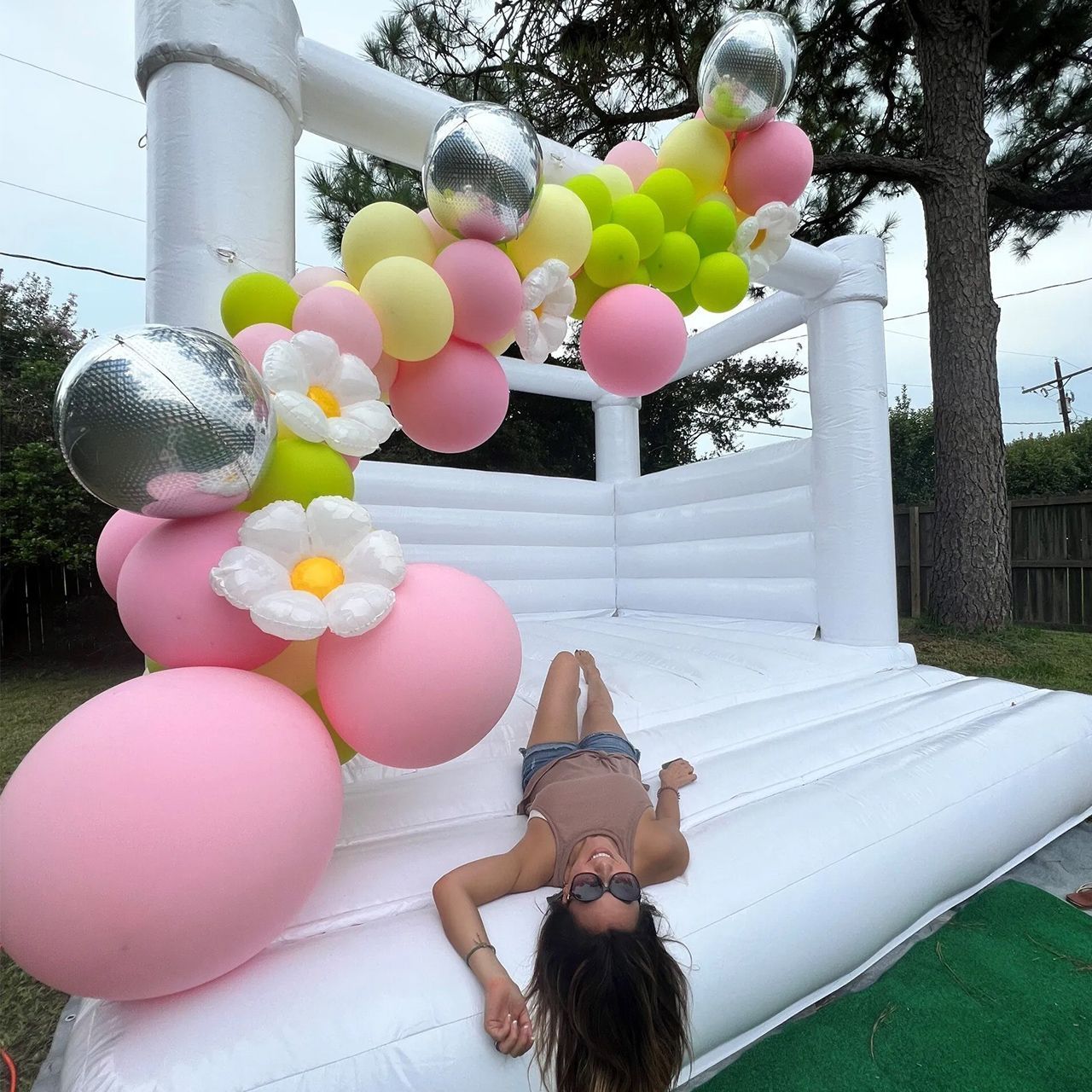 A young person smiles lying on a white bounce house with a balloon arch.