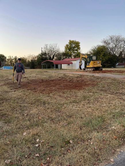 A man is walking in a field with a shovel and a bulldozer in the background.