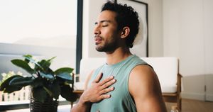 Man sitting indoors with eyes closed, hand on chest, in a calm breathing pose near a plant and window.