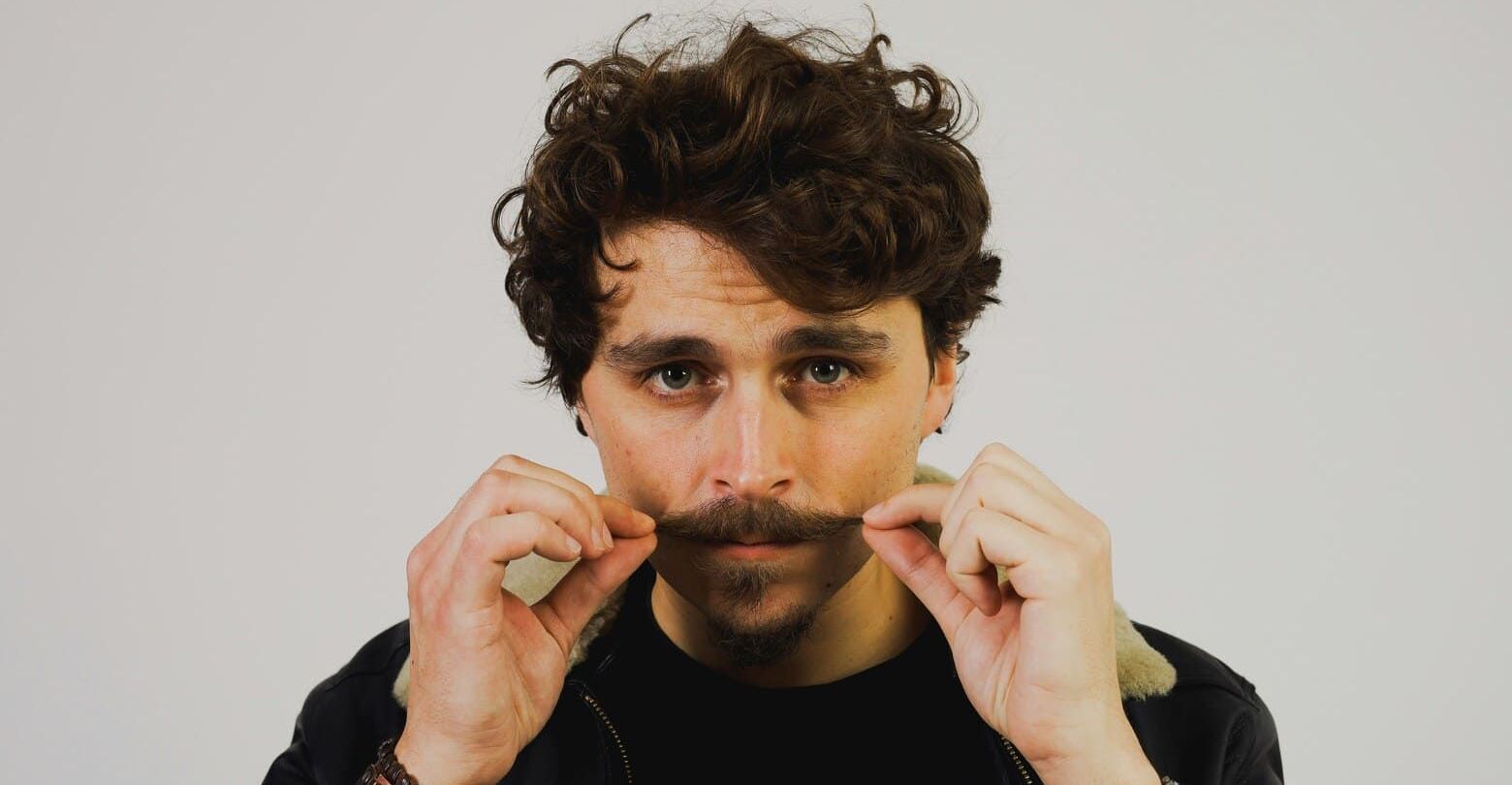 A man with curly hair is holding his mustache in front of his face.