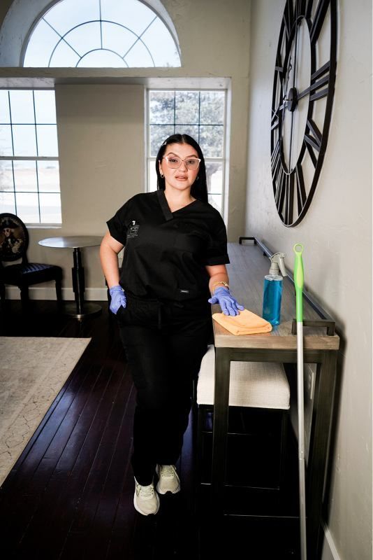 Person in black uniform and blue gloves stands by a cleaning cart in a bright room with large windows.
