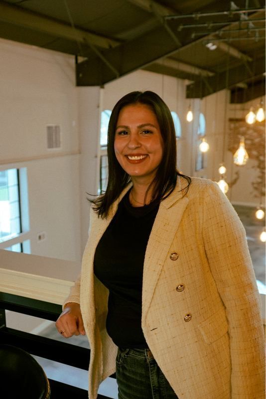 Woman smiling on an indoor balcony, wearing a black top and beige cardigan.