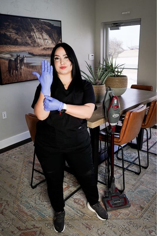 Person in black clothes wearing blue gloves, standing in a dining room with a table, chairs, and plant.