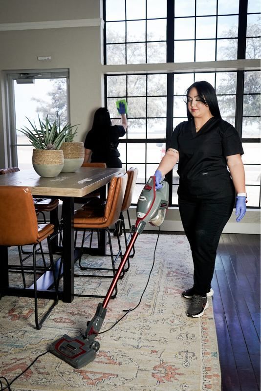 Person vacuuming a rug in a bright dining room with large windows.