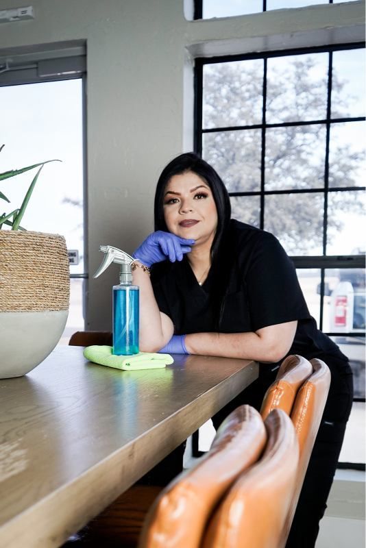 Woman seated at a wooden table by a window, resting her chin on her hand beside a blue bottle and green notebook