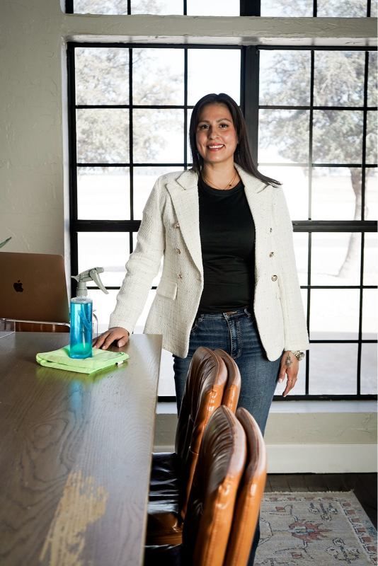 Woman standing in an office beside a table, wearing a white blazer and smiling in front of large windows