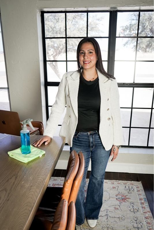 Woman standing in a bright room beside a desk and chair, wearing a white blazer and jeans.