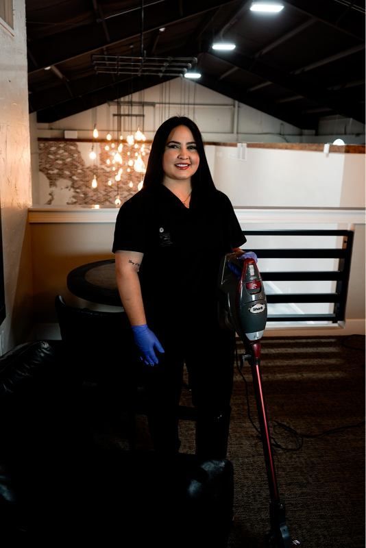 Woman in black uniform with blue gloves stands indoors beside a vacuum cleaner and cart.