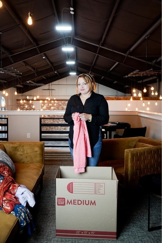 Person holding a pink jacket beside a cardboard box in a loft-style room with sofas and string lights