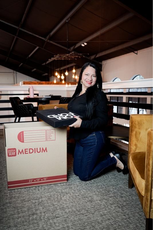 Woman kneeling beside a large box in an attic, holding a tray, with exposed beams and warm lights.