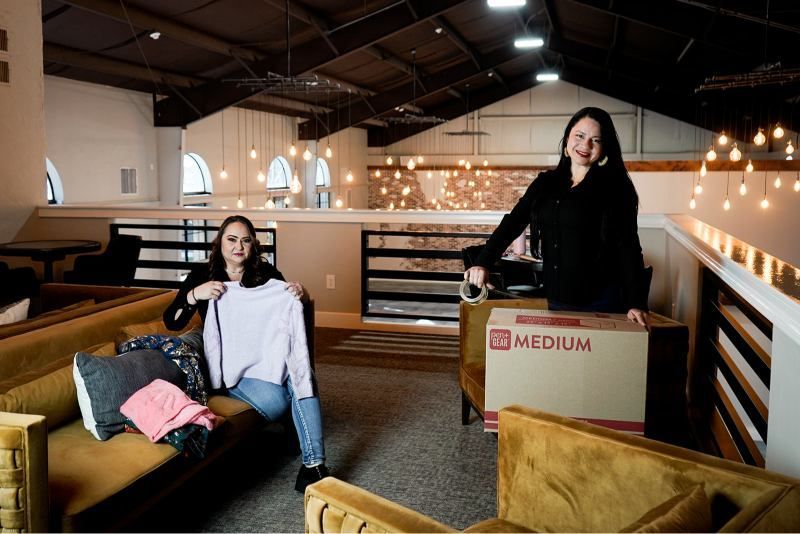 Two people with boxes and a couch in a loft-style room with string lights