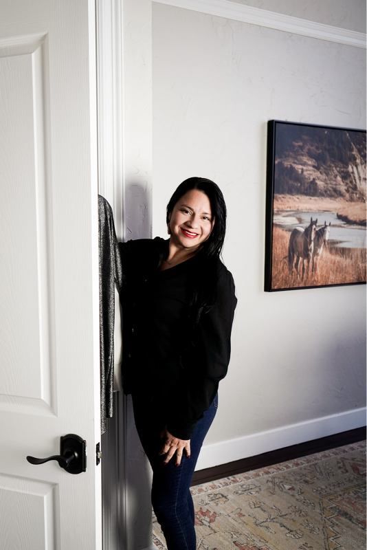 Woman peeking from behind a doorway in a hallway, smiling, with framed wall art and a rug nearby.
