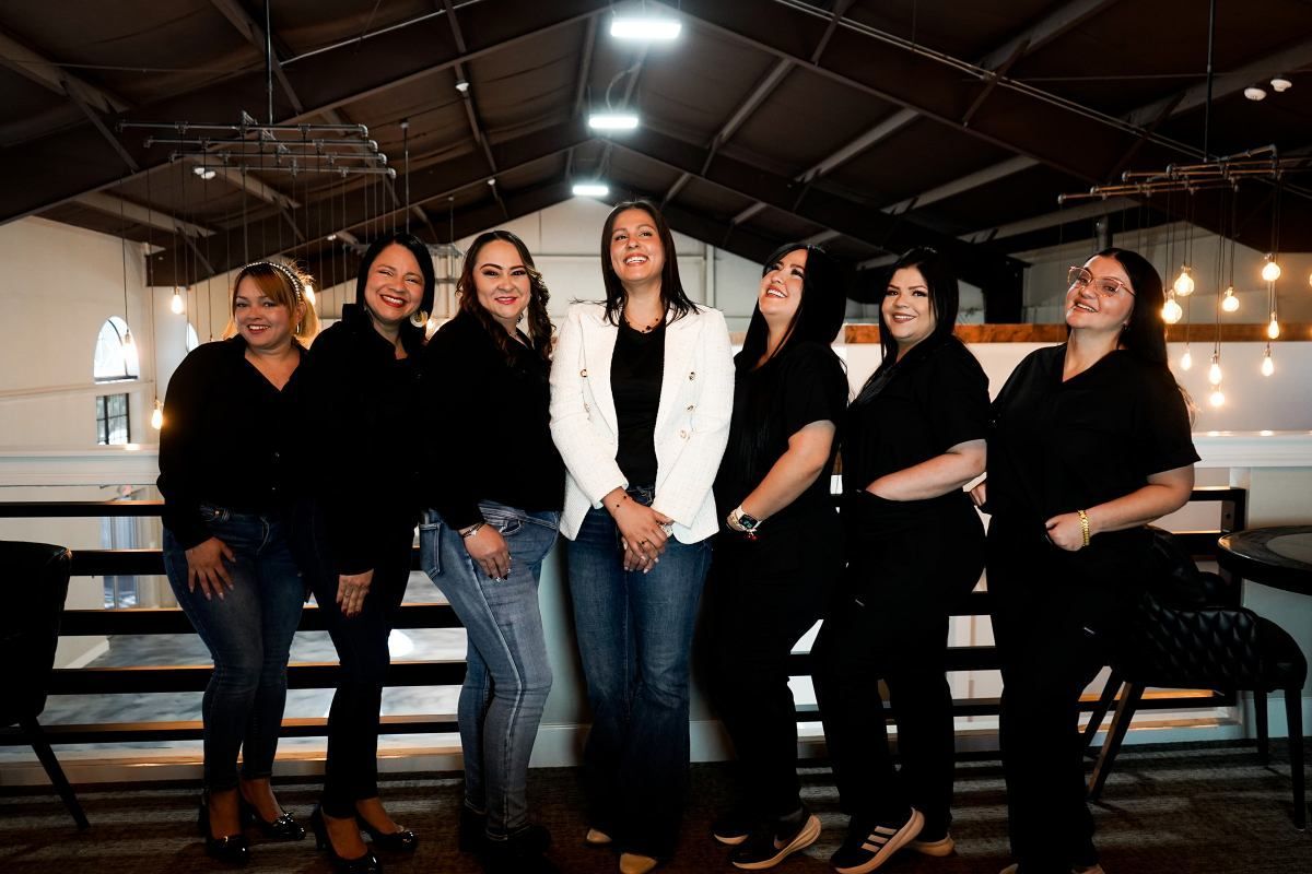 Group of six people posing together indoors under a barn-style roof, dressed mostly in black with one white jacket.