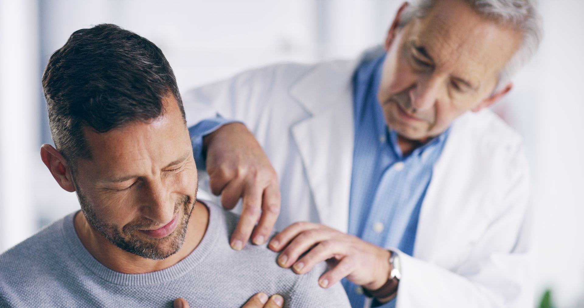 A doctor in a white coat comforts a man in a grey sweater; both are indoors. The man holds his chest, appearing distressed.