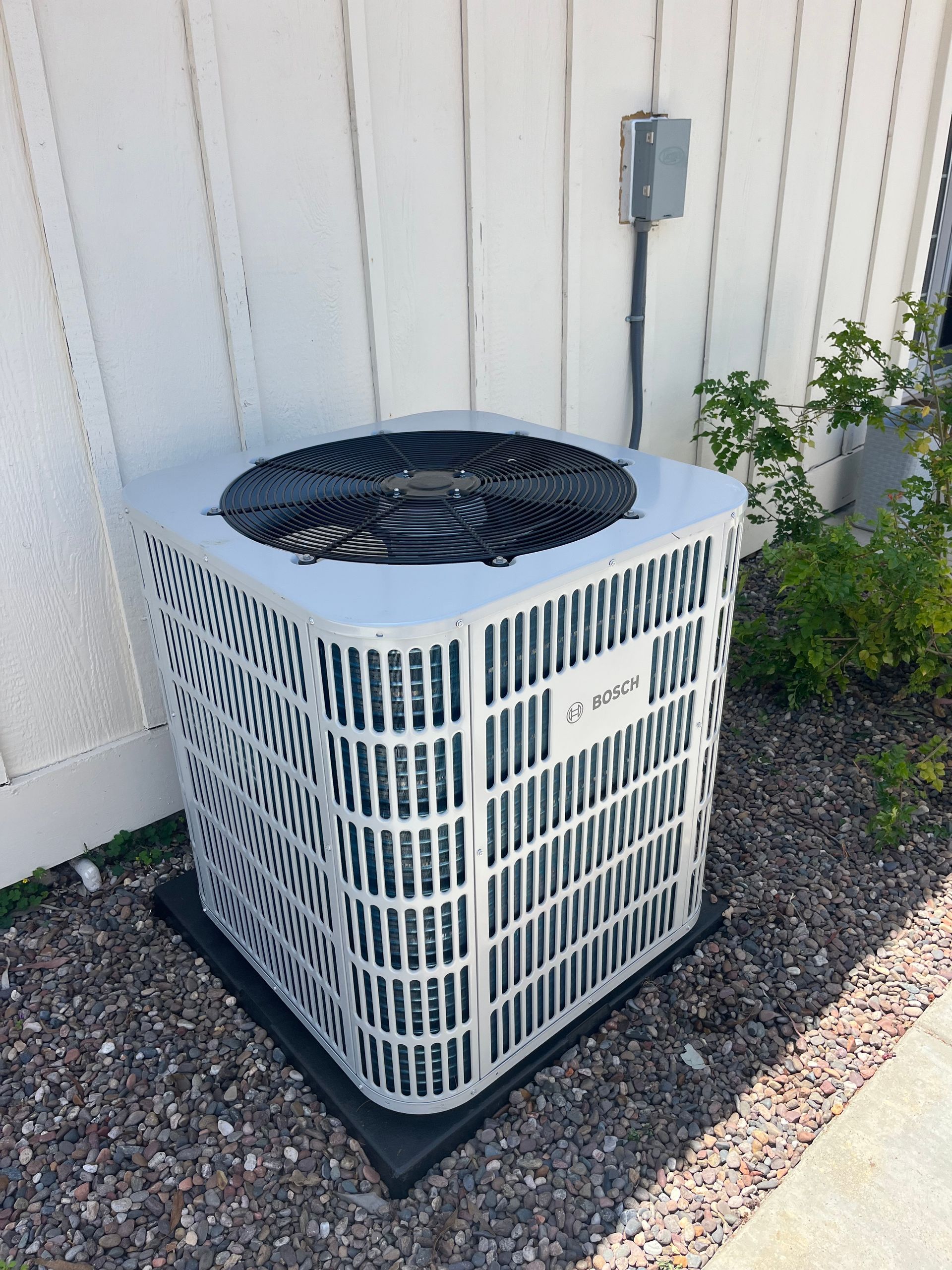 Air conditioner unit outside against a white wall with landscaping.
