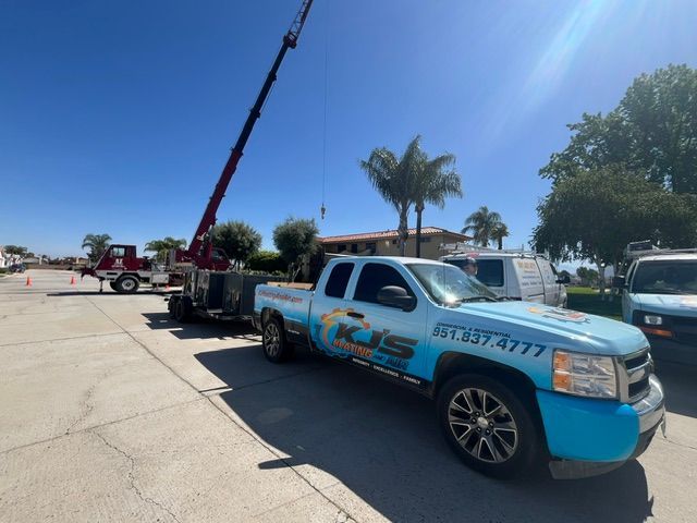 A crane lifting something, with a KJS truck parked nearby in front of a building on a sunny day.